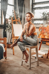 Glamour female painter with collected blond hairs sitting on chair and holding paintbrushes poses in her professional studio.