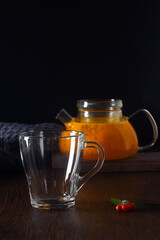 Hot tea with sea buckthorn in a glass pot and empty glass on the dark wooden table, knitting blanket near. Healthy drink, cozy winter night. Vertical orientation, copy space.