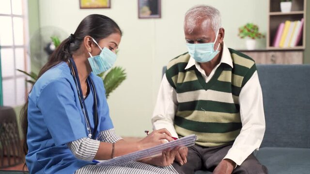 Young doctor or nurse writing prescription during home visiting to sick elder man while both worn face mask due to coronavirus covid-19 pandemic