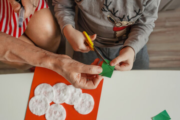 A Little boy with grandmother make Christmas craft, greeting card. Top view boy cuts a triangle out of paper with scissors.