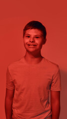 Obraz premium Portrait of joyful teenaged disabled boy with Down syndrome smiling at camera while posing isolated over red light background
