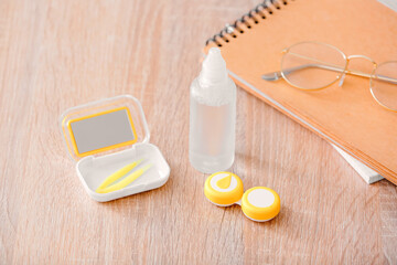 Container with contact lenses, solution, eyeglasses and tweezers on wooden table