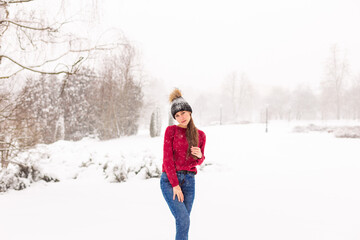 Fototapeta na wymiar A young beautiful girl in a red sweater stands among the snowdrifts in the forest. Woman in red in a park winter when it snows.