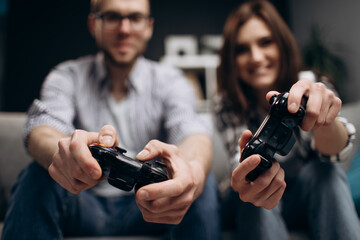 Blur background of young couple resting on grey couch and using joysticks for playing video games. Focus on hands and remote controllers. Concept of entertainment.