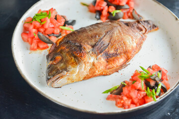 Fried fish with fresh herbs, tomatoes and olives served on a white plate over black rustic wooden table background.