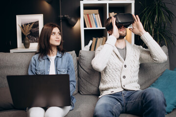Attractive man and woman relaxing together at home and using VR headset with modern laptop. Young couple playing virtual games while sitting on couch.