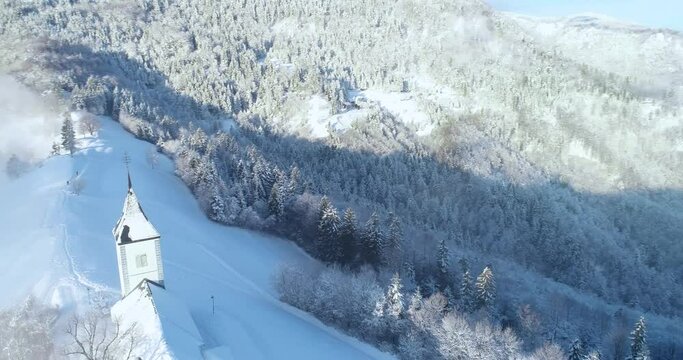 Aerial drone flying over famous Jamnik church, Slovenia. Landscape covered with white snow. Fairy tale like nature and small catholic church on hilltop. Dynamic moving, parallax