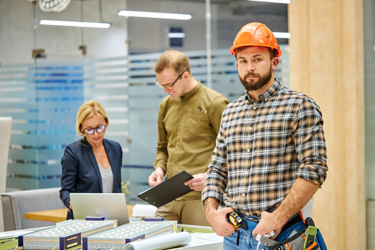 portrait of confident engineer male in safety helmet, young caucasian man posing at camera, they work on planning building project, using layout. colleagues in the background