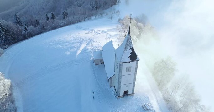 Aerial drone flying over famous Jamnik church, Slovenia. Landscape covered with white snow. Fairy tale like nature and small catholic church on hilltop. Forward moving, tilt down