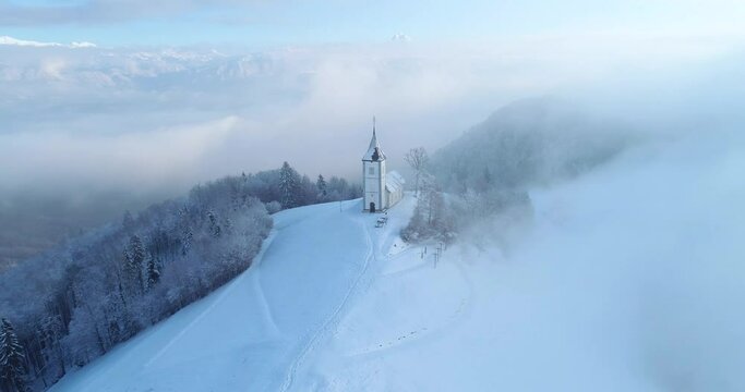 Aerial drone flying over famous Jamnik church, Slovenia. Landscape covered with white snow. Fairy tale like nature and small catholic church on hilltop. Alps mountains in the distance