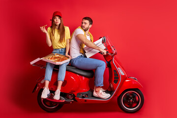 Fototapeta na wymiar funny redhead woman rides with courier boyfriend delivering food orders on motorcycle, female is eating pizza sitting behind man, isolated red background