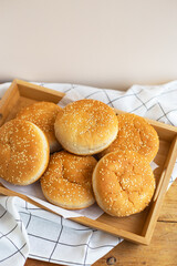Freshly baked hamburger buns lie on a wooden tray on the table with a white napkin. Yellow flavored pastries with white sesame seeds.