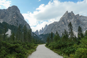 Fototapeta na wymiar A gravel road through a forest, leading to high and sharp Italian Dolomites. The road is very curvy. There are thick clouds above the mountain peaks. Idyllic landscape. Isolated and remote place