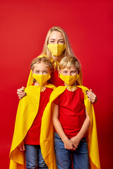 fight for health, defeating the disease. blonde caucasian mother stand with son and daughter in superhero cloaks and medical masks isolated over red background
