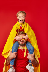 surprised super girl sits on neck of father closing his eyes, superhero man hold child and smiles....