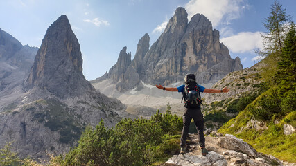 Fototapeta premium A man with big hiking backpack hiking in high Italian Dolomites. There are many sharp peaks behind. He is spreading his arms in the gesture of freedom. There are a few trees around. Sunny day. Outdoor
