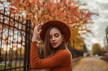 Young elegant, fashionable woman wearing stylish orange hat, trendy round earrings, posing in street. Outdoor close up autumn portrait. Copy, empty space for text