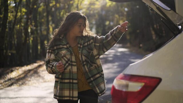 Happy young woman in checkered jacket opens trunk of white automobile to take map and thermocup on empty rural road in picturesque autumn forest