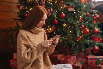 Side view of charming young woman getting Christmas greeting card from her beloved on background of xmas tree at cozy living room with festive interior. Surprised lady pleasure reading letter at home.