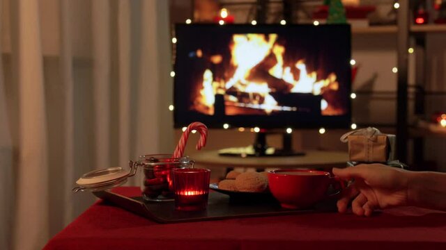 winter holidays and hygge concept - hand placing cup of coffee or hot chocolate to table with ginger cookies, candle, candy canes and gift boxes over tv monitor used as fireplace at cozy home