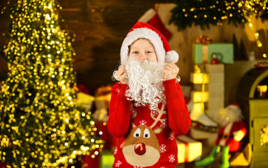December tradition. Fluffy beard. Child with white long fake beard near christmas tree. Merry christmas. Happy childhood concept. Kid wear santa hat and christmas sweater. Santa brought me gifts