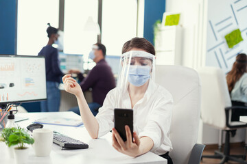 Businesswoman having a corporate video conference using smartphone wearing face mask. Colleagues working respecting social distance during global pandemic with coronavirus.