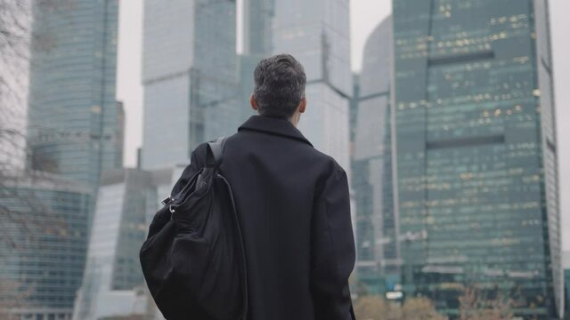 Man in black coat going towards business city skyscrapers in the evening, slow motion, lights in the windows. Gimbal shot of young businessman, no face, with bag
