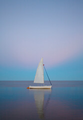 A white boat in the middle of a calmed sea with a pink and blue sunset background. The boat is a sailboat in a peaceful place.
