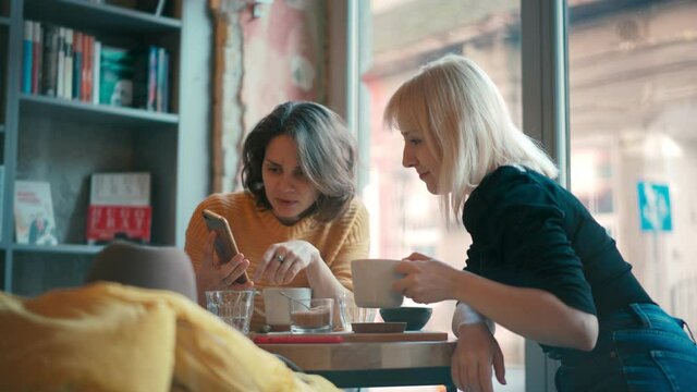 Handheld cinematic shot of two young women sitting at the hipster cafe, having a conversation and showing each other something on their phones.