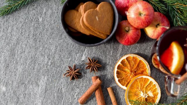 christmas and seasonal treats concept - snow falling over glass of hot mulled wine with orange slice, gingerbread cookies, apples and fir branch on grey background