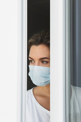 Scared woman wearing prevention mask looking outside through the window