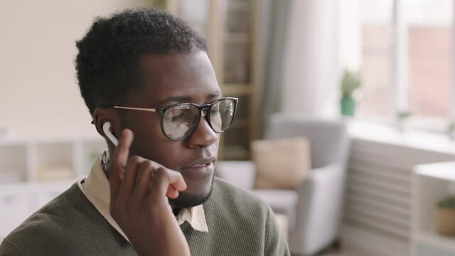 Medium shot of young African-American businessman wearing eyeglasses and wireless earphones sitting at workplace and receiving phone call via headphones