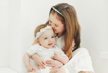 Portrait of cute little girl child with her sister baby sitting in white room at home near window