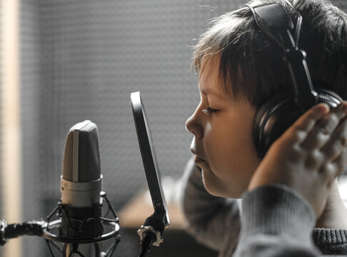 Boy in check shirt and headphone is singing or talking into microphone with pop filter in voice recording studio. Young singer recording his voice for phonogram performance for entertainment.
