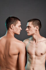 Young caucasian half naked twin brothers looking at each other while posing in studio, standing isolated over grey background