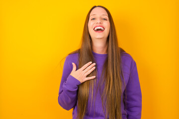 Young beautiful Caucasian woman wearing purple sweater against yellow wall smiles toothily cannot believe eyes expresses good emotions and surprisement