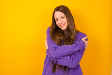 Charming pleased Young beautiful Caucasian woman wearing purple sweater against yellow wall embraces own body, pleasantly feels comfortable poses. Tenderness and self esteem concept