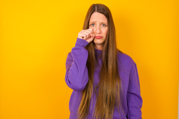 Disappointed dejected Young beautiful Caucasian woman wearing purple sweater against yellow wall wipes tears stands stressed with gloomy expression. Negative emotion