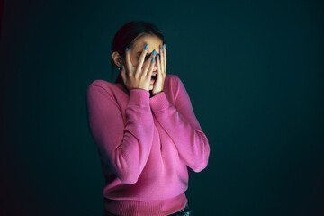 Hiding face. Portrait of young crazy scared and shocked caucasian woman isolated on dark background. Copyspace for ad. Bright facial expression, human emotions concept. Looking horror on TV, cinema.
