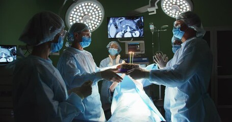 Happy team of professional doctors in operating room in hospital under bright lamp congratulating surgeon wishing happy birthday, giving birthday cake, celebrating clapping hands. Surgeons and nurses - Powered by Adobe