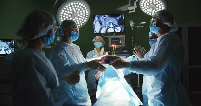 Happy team of professional doctors in operating room in hospital under bright lamp congratulating surgeon wishing happy birthday, giving birthday cake, celebrating clapping hands. Surgeons and nurses