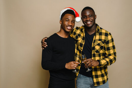 Happy family portrait on Christmas, gay male couple on beige background. A pair of black men in love clink glasses of champagne. Cheers. Celebrate Christmas and New Year