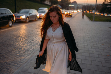 red-haired young woman in a white dress in autumn runs down the street barefoot carries shoes in...