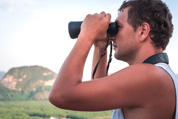 A young man in a white T-shirt looks from the top into the distance through binoculars against the background of the sky and mountains close-up