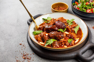 Tagine lamb simmered in spices, with dates and almonds, salad with tomatoes, coriander and parsley on grey background with copy space.