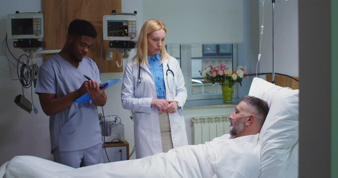 Caucasian middle-aged male patient lying on bed in hospital suffering from coronavirus and speaking with female doctor while African American man assistant writing down symptoms. Healthcare concept