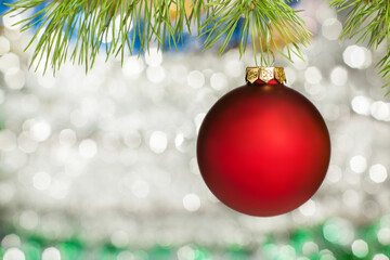 Christmas decoration - a red matte ball hanging on a branch of a Christmas tree. In the background, blurry flickering lights of a garland. Suitable for banner making, postcards and any New Year design