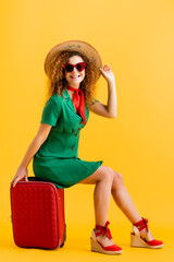 full length of cheerful woman in straw hat, sunglasses and dress sitting on luggage on yellow