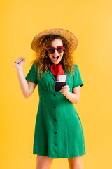 amazed woman in straw hat, sunglasses and dress holding passport on yellow