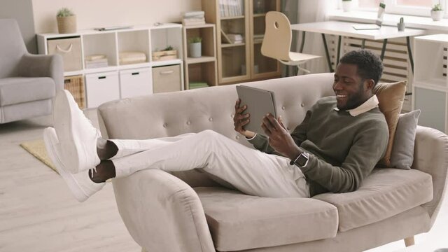 Wide shot of young African-American businessman wearing casual clothes lying on couch in living room with tablet in his hand and having video chat with colleague